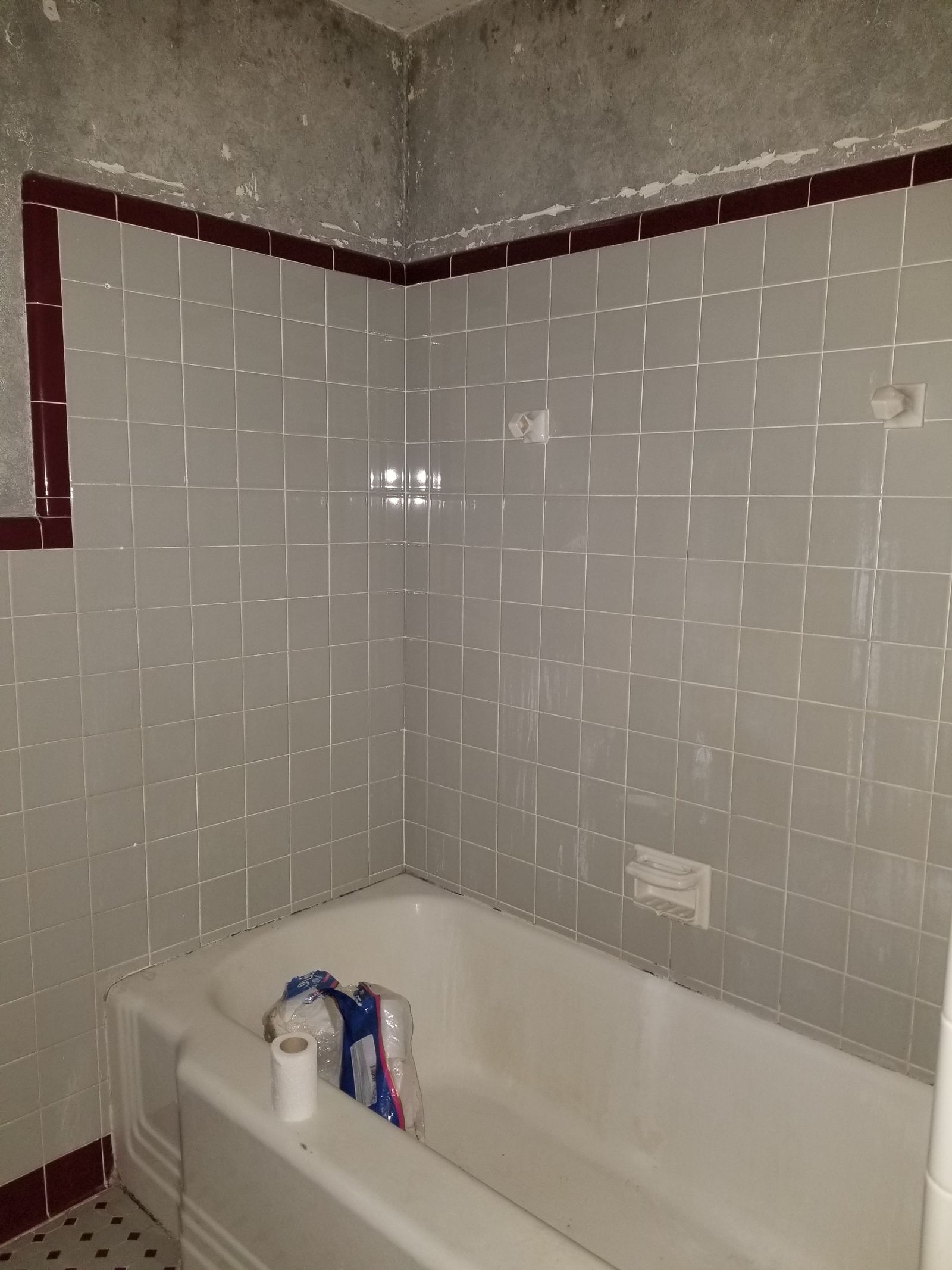 Original tub surround with aging gray and burgundy tile, peeling wall paint, and worn caulk lines