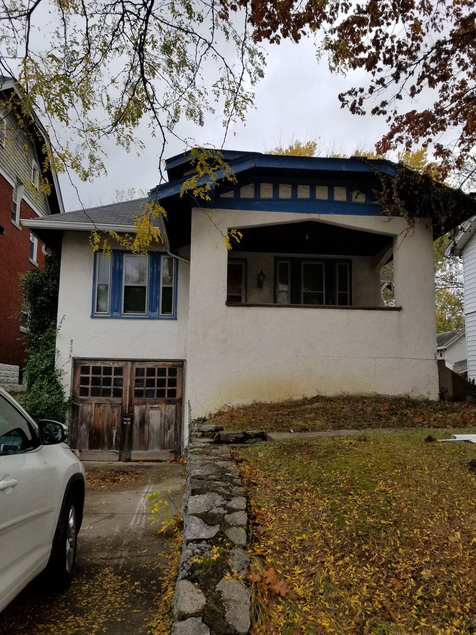 Front of the bungalow before renovation, with peeling blue trim, weathered wooden garage doors, and an overgrown front slope