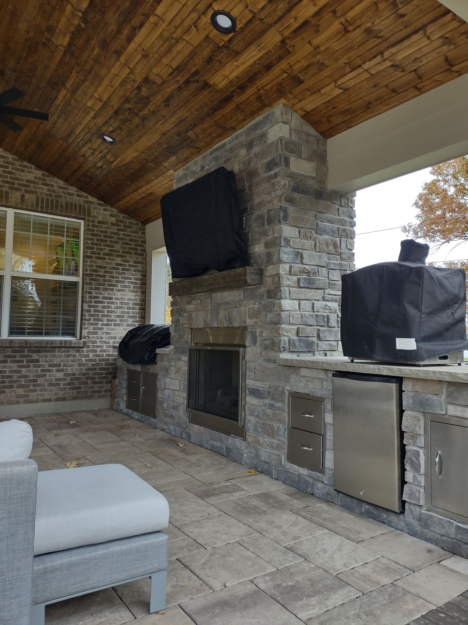 Covered outdoor kitchen with stone fireplace, mounted TV, and stainless appliances