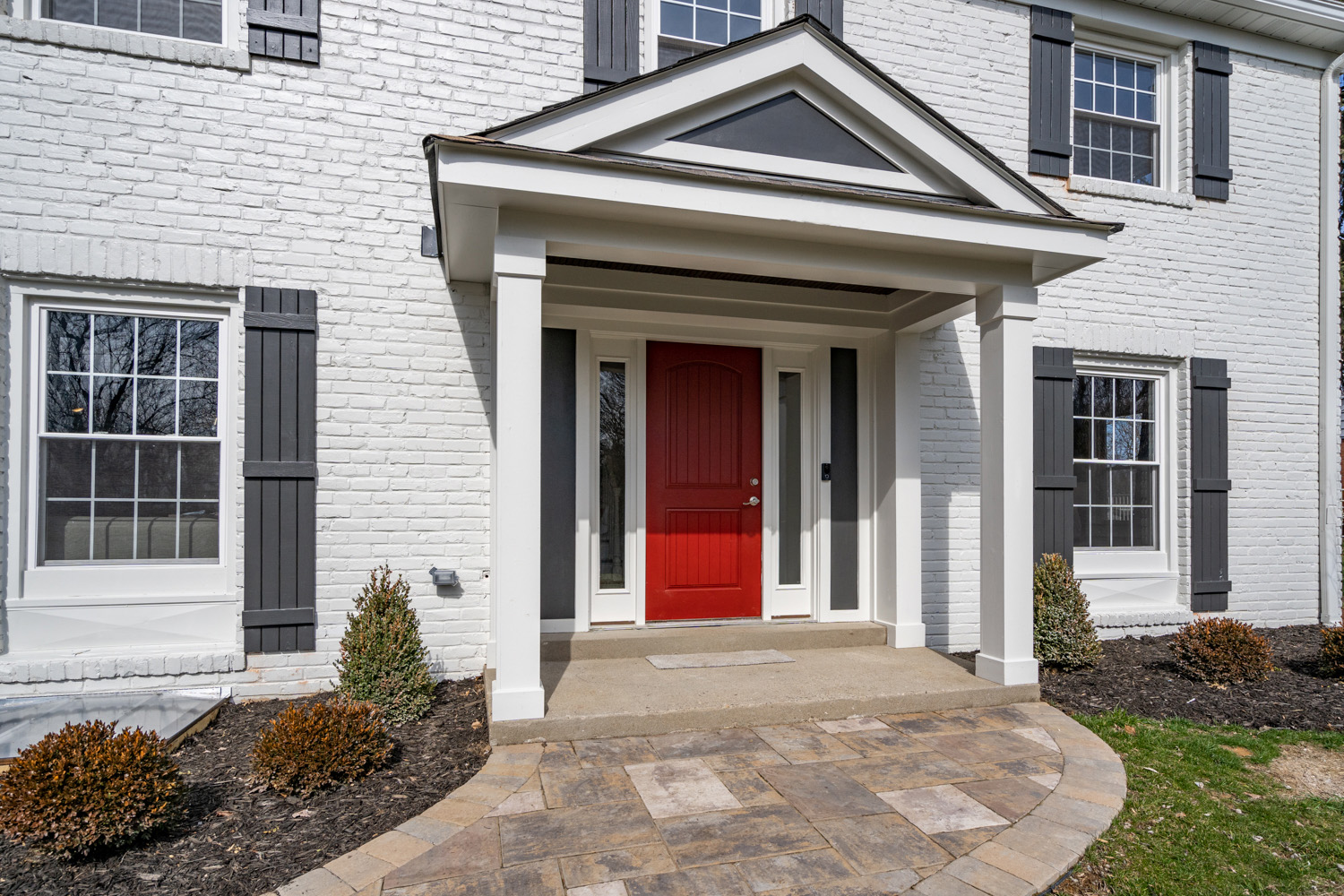White brick colonial with columned entry portico, red front door, and paver walkway